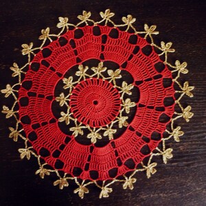 Può includere: Tovaglietta all'uncinetto rossa con bordo dorato. La tovaglietta ha un design circolare con un cerchio più piccolo al centro.