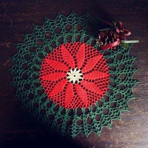 Könnte beinhalten: Handgefertigte, gehäkelte Tischdecke im Poinsettia-Design. Die Mitte ist rot mit einer cremefarbenen Mitte, umgeben von einem grünen, durchbrochenen Rand. Ein Tannenzapfen mit rotem Band und grünem Zweig liegt am Rand der Tischdecke.