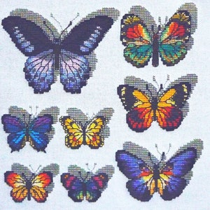 May include: A collection of seven cross-stitch butterflies in various sizes and colors. The butterflies feature detailed wing patterns in shades of blue, purple, orange, yellow, and green. The background is a white fabric.