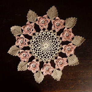 Könnte beinhalten: Eine zarte gehäkelte Tischdecke mit einem runden Zentrum und einem Sternmuster. Die Tischdecke ist aus beigefarbenem und rosafarbenem Garn gefertigt und hat ein florales Design.