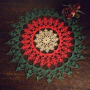 Può includere: Un centrino all'uncinetto rotondo di colore verde, rosso e oro. Il centrino ha un centro con un motivo a stella ed è circondato da un anello rosso e poi da un anello verde.