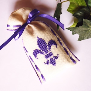 Può includere: Un piccolo sacchetto di stoffa beige con un disegno di giglio viola. Il sacchetto è legato con un nastro viola e presenta dettagli viola sui lati. Probabilmente riempito con pot-pourri o altri articoli profumati.