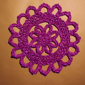 Può includere: Un centrino all'uncinetto rosa con una finitura scintillante. Il centrino ha una forma circolare con un disegno floreale al centro.