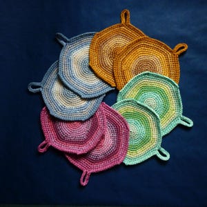 Può includere: Una collezione di presine fatte a mano, all'uncinetto, in vari colori, tra cui blu, oro, rosa e verde. Ogni presina ha un anello per appenderla e una forma esagonale. Le presine sono disposte in un motivo circolare su una superficie blu scuro.