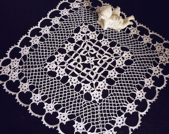 May include: White crocheted doily with a delicate floral pattern and a heart-shaped border. The doily is laid flat on a dark surface.