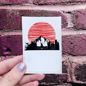 May include: A sticker with a black silhouette of a city skyline with a mountain in the center, set against a red sun with white stripes.