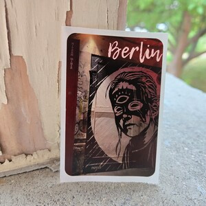May include: A sticker with a black and white illustration of a person wearing a mask. The background is a red and black brick wall with graffiti. The sticker says "Berlin" in pink letters.