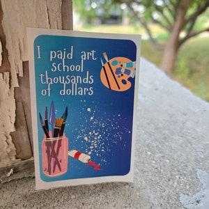 Könnte beinhalten: Ein blauer und weißer Aufkleber mit dem Text "Ich habe Tausende von Dollar für die Kunstschule bezahlt". Der Aufkleber zeigt eine Palette, Farbtuben und Pinsel.