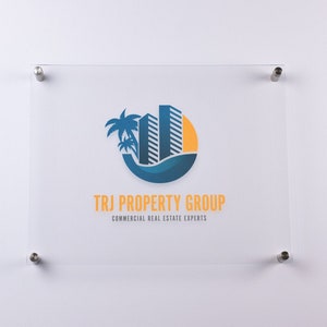 May include: A white sign with a blue and yellow logo of a palm tree and two buildings in a circle. The text "TRJ PROPERTY GROUP" and "COMMERCIAL REAL ESTATE EXPERTS" is below the logo.