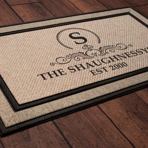 May include: Rectangular doormat with a beige woven texture and a black border. The mat features the name "THE SHAUGHNESSYS" with "EST 2000" below, and a stylized initial "S" in a circle with decorative flourishes.