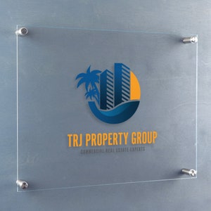 May include: A glass sign with a blue and yellow logo of a building with palm trees and a sun. The text "TRJ PROPERTY GROUP" is below the logo, with the tagline "COMMERCIAL REAL ESTATE EXPERTS" underneath.