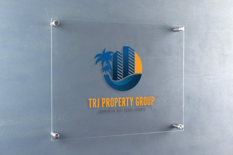 May include: A glass sign with a blue and yellow logo of a building with palm trees and a sun. The text "TRJ PROPERTY GROUP" is below the logo, with the tagline "COMMERCIAL REAL ESTATE EXPERTS" underneath.