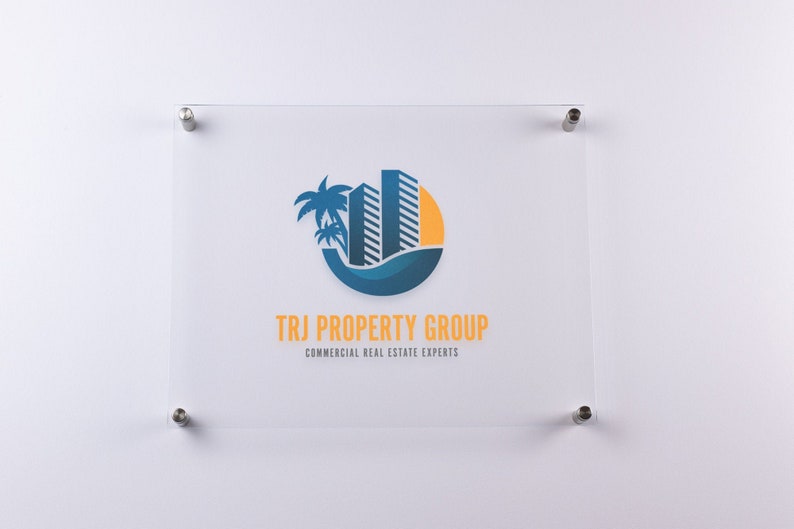 May include: A white sign with a blue and yellow logo of a palm tree and two buildings in a circle. The text "TRJ PROPERTY GROUP" and "COMMERCIAL REAL ESTATE EXPERTS" is below the logo.