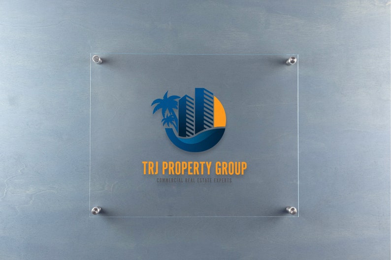 May include: A clear glass sign with a blue and yellow logo of a palm tree and buildings with the text "TRJ PROPERTY GROUP" and the tagline "COMMERCIAL REAL ESTATE EXPERTS".