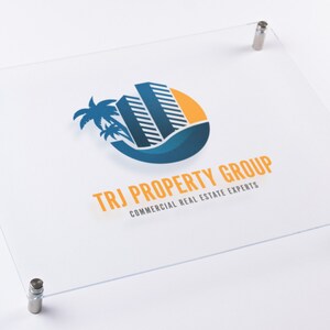 May include: A clear acrylic sign with rounded corners and silver standoffs. The sign features a logo with a blue and yellow stylized image of a building with palm trees and the text "TRJ PROPERTY GROUP COMMERCIAL REAL ESTATE EXPERTS".