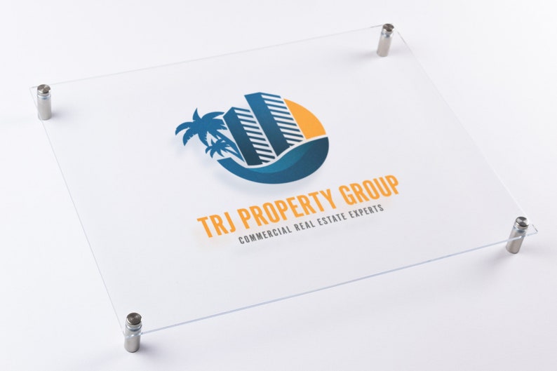 May include: A clear acrylic sign with rounded corners and silver standoffs. The sign features a logo with a blue and yellow stylized image of a building with palm trees and the text "TRJ PROPERTY GROUP COMMERCIAL REAL ESTATE EXPERTS".