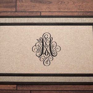 May include: Rectangular doormat with a woven beige center and a dark brown border. The center features a black, ornate monogram design. The mat is designed for placement at an entryway.