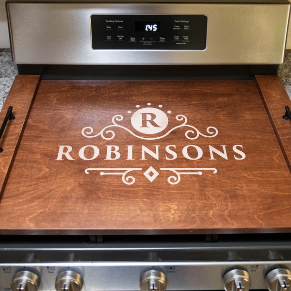 Könnte beinhalten: Ein Herdplattenabdeckung aus Holz mit einem personalisierten Design. Die Abdeckung ist braun mit einem weißen Monogramm und dem Namen "ROBINSONS" in einer dekorativen Schrift.