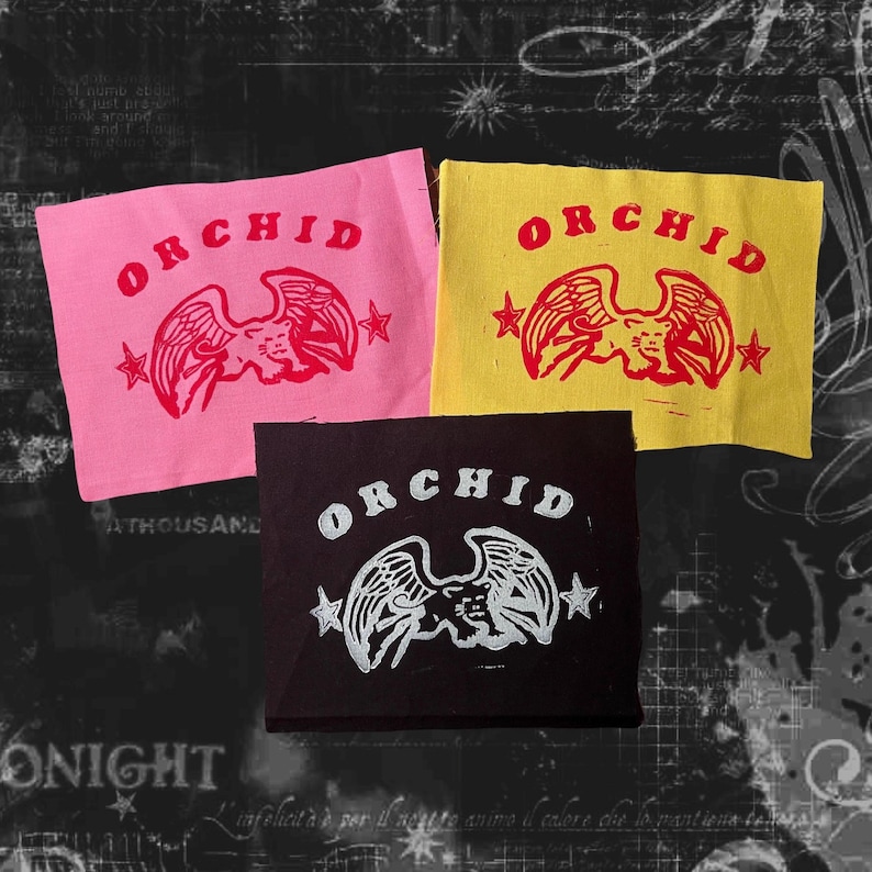May include: Three fabric patches with the word "ORCHID" printed in white, red, and black lettering. Each patch features a stylised eagle design with wings spread, printed in red, white, and black. The patches are on a dark background.
