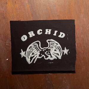 May include: Black fabric patch with a white graphic of a winged creature with the word "ORCHID" above it.
