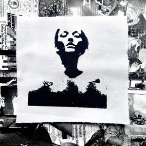 以下が含まれることがあります： 正方形の白い布パッチ。人の顔と肩の黒いシルエットが特徴です。画像は様式化された肖像画です。背景は、新聞の切り抜きやその他の印刷物のコラージュです。