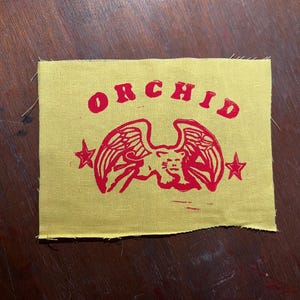 May include: A yellow fabric patch with a red printed design. The design features a winged creature with a star on each side and the word "ORCHID" above it.