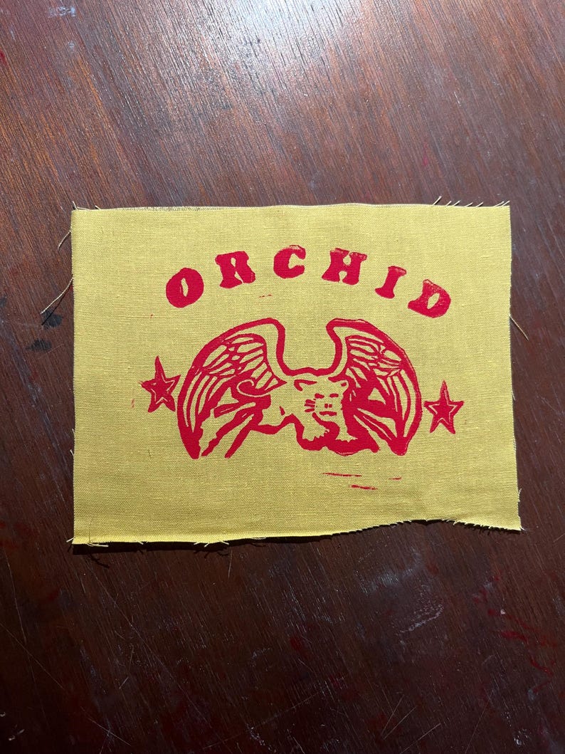 May include: A yellow fabric patch with a red printed design. The design features a winged creature with a star on each side and the word "ORCHID" above it.