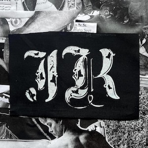 以下が含まれることがあります： 黒い長方形のパッチ。白い文字「JR」が装飾的なゴシック様式のフォントで描かれています。文字には装飾的な装飾と詳細があります。パッチは白黒の画像が背景にあります。