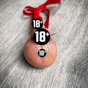 Puede incluir: Un adorno novedoso con forma de testículo, con el número 18+ mostrado en tres círculos negros. El adorno es de color rosa claro, con una cinta roja atada en la parte superior. El fondo es una superficie gris texturizada.