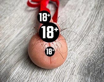 May include: A novelty ornament shaped like a testicle, with the number 18+ displayed on three black circles. The ornament is a light pink color, with a red ribbon tied at the top. The background is a gray, textured surface.