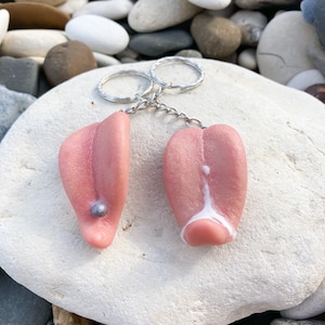 Op de afbeelding: Twee roze tongvormige sleutelhangers met een zilveren ketting. Eén sleutelhanger heeft een zilveren kraal op de tong. De andere sleutelhanger heeft een witte substantie op de tong.