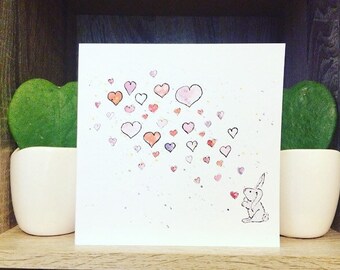 Puede incluir: Una pintura de acuarela de un conejo blanco soplando corazones rosas, rojos y morados. El fondo es blanco con un ligero lavado de acuarela azul.