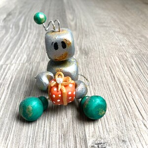 May include: A silver robot figurine holding a small orange gift box with white polka dots. The robot has green and gold accents and a wire antenna.