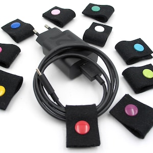 Può includere: Un set di dieci organizzatori di cavi con bottoni di diversi colori. Gli organizzatori sono in feltro nero e sono progettati per mantenere i cavi e i fili in ordine.