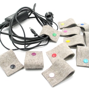 Può includere: Un set di dieci organizzatori per cavi in feltro grigio con bottoni colorati. Ogni organizzatore ha un bottone di un colore diverso, tra cui rosso, blu, verde, giallo, rosa, viola, nero e bianco.