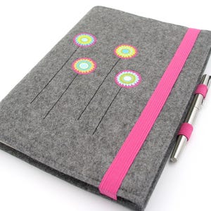 Könnte beinhalten: Ein grauer Notizbuchumschlag aus Filz mit vier gestickten Blumenmotiven in Pink, Gelb und Blau. Ein leuchtend pinkfarbenes Gummiband hält den Umschlag zusammen, während ein silberner Metallkugelschreiber in einer passenden pinken Schlaufe steckt.