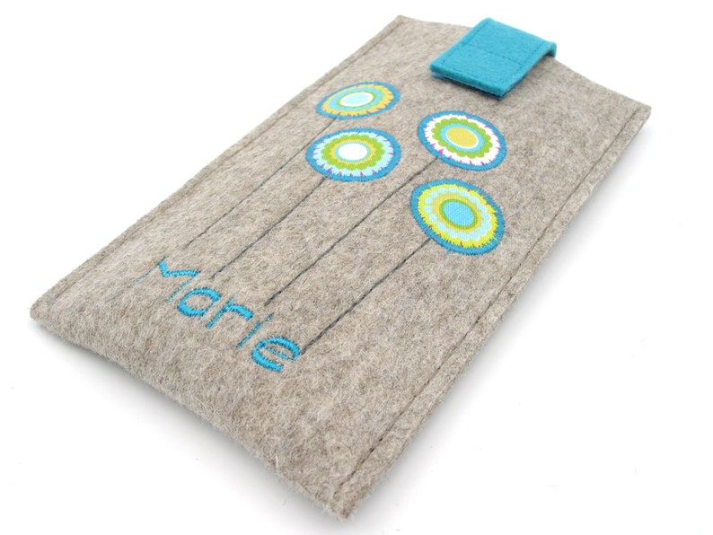 May include: A gray felt phone case with a blue flap closure. The case has a blue and green floral design and the name "Marie" embroidered on the front.