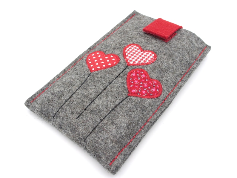 May include: Grey felt phone case with a red felt flap closure. Three red felt hearts with different patterns are stitched on the front of the case.