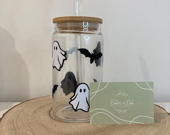 May include: A clear glass tumbler with a bamboo lid and straw, decorated with white ghost and black bat decals. A business card with the text "Ember & Oak Crafts" is next to the tumbler. The tumbler is on a wooden surface.
