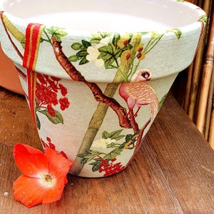 Puede incluir: Una maceta de terracota con fondo azul claro y un patrón floral con pájaros y bayas rojas. La maceta está decorada con una cinta roja y dorada.