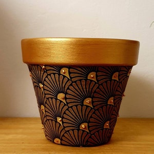 Puede incluir: Una maceta de terracota pintada de oro con un patrón geométrico negro y marrón. La maceta tiene detalles dorados alrededor del borde y en todo el patrón.