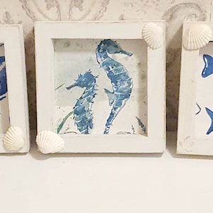 Peut inclure: Trois tableaux carrés encadrés de blanc. Chacun présente un motif aquarelle bleu et blanc : un homard, des hippocampes et deux poissons. Des coquillages ornent les coins de chaque cadre, soulignant le thème côtier.