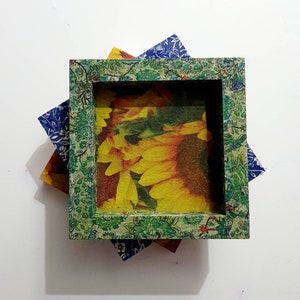 Puede incluir: Un marco de fotos cuadrado de madera con un patrón floral verde. El marco está superpuesto sobre otras tres piezas cuadradas de tela con patrones florales en azul, amarillo y rojo.