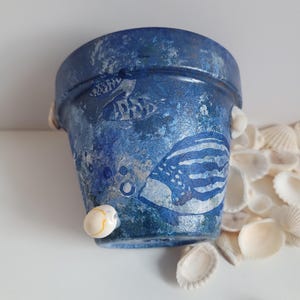 Könnte beinhalten: Ein blauer Terrakotta-Pflanztopf mit einem weißen, bemalten Design aus Fischen und Muscheln. Der Topf hat einen Used-Look und ist mit kleinen weißen Muscheln verziert.