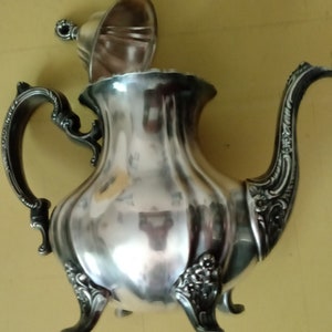 May include: A silver teapot with a lid and ornate details. The teapot has a round body with a spout and handle. The teapot is sitting on a yellow surface.