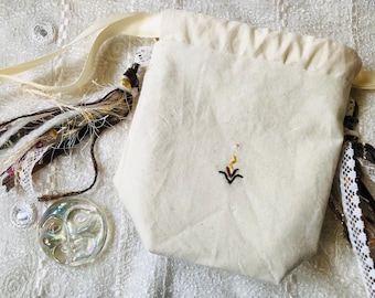 May include: A cream-colored drawstring bag with a small embroidered design. The bag is accompanied by decorative tassels with feathers, lace, and beads, and a clear glass moon-shaped object. The items are arranged on a textured white surface.