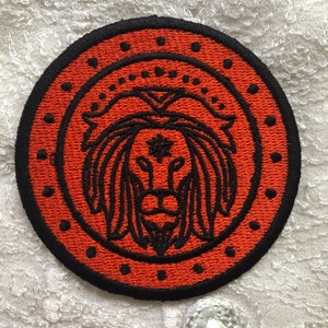 May include: A round, orange embroidered patch with a black outline. The patch features a lion's head with a star in the centre.