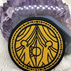 Puede incluir: Parche bordado redondo con borde negro y diseño central dorado. El diseño presenta un emblema estilizado con figuras y líneas simétricas. El parche es adecuado para ropa o accesorios.