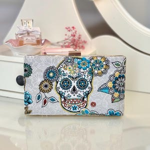 Peut inclure: Une pochette rectangulaire avec un motif de tête de mort mexicaine. La pochette présente un fond blanc avec des détails floraux et de crâne colorés en bleu, jaune et rouge. Le fermoir est de couleur dorée.