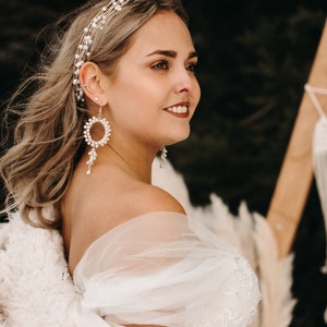 Könnte beinhalten: Eine Braut in einem weißen, schulterfreien Brautkleid mit einem Perlendiadem und Perlenohrringen. Die Braut lächelt und blickt nach rechts.